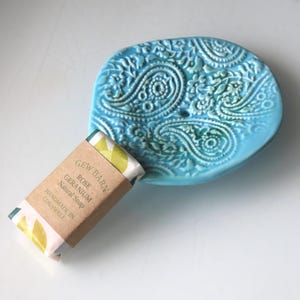 May include: A blue ceramic soap dish with a paisley pattern. The dish is filled with a bar of soap wrapped in brown paper with the text "GEW BARON ROSE GERANIUM Natural Soap HANDMADE IN CORNWALL".