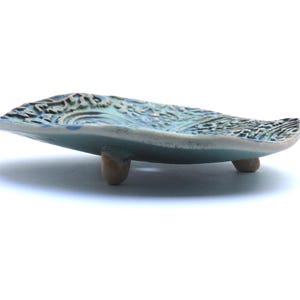 May include: A small, teal blue ceramic dish with a decorative, textured surface. The dish has three small, rounded feet.