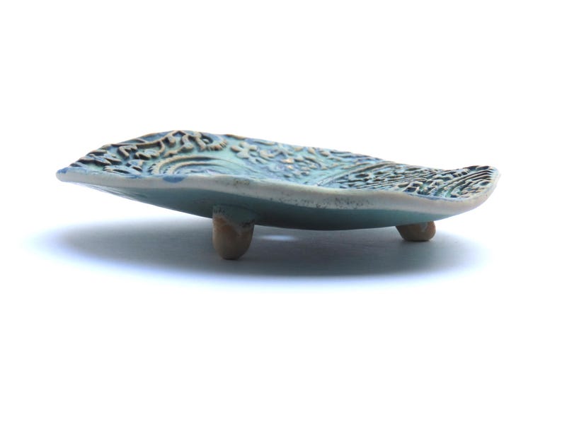 May include: A small, teal blue ceramic dish with a decorative, textured surface. The dish has three small, rounded feet.