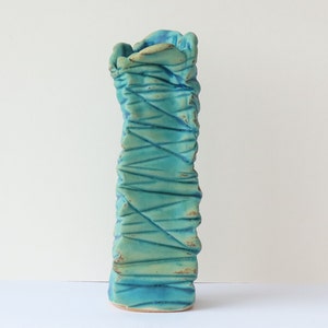 May include: A tall, turquoise ceramic vase with a textured, organic shape. The vase has a rough, uneven surface and a slightly flared opening.