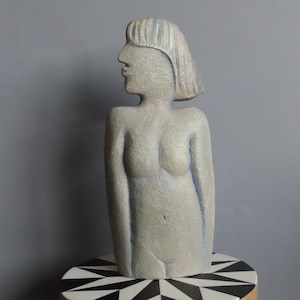 Puede incluir: Una escultura de piedra gris de una mujer con un corte de pelo corto. La escultura está de pie sobre una superficie con un patrón blanco y negro.