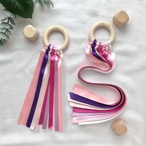 Puede incluir: Dos sonajeros para beb&eacute;s con aros de madera y cintas de raso de colores en tonos rosa, morado y blanco. Cada sonajero tiene el texto "Pishu Baby" en el aro de madera. Bloques de madera dispersos.