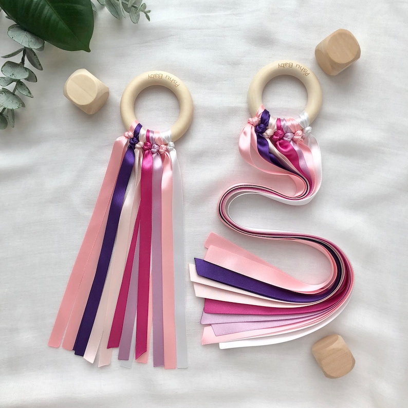 Puede incluir: Dos sonajeros para beb&eacute;s con aros de madera y cintas de raso de colores en tonos rosa, morado y blanco. Cada sonajero tiene el texto "Pishu Baby" en el aro de madera. Bloques de madera dispersos.