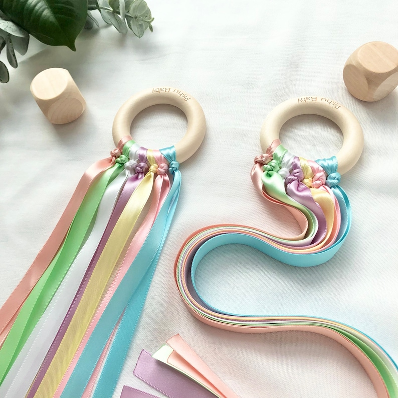 Puede incluir: Dos juguetes para beb&eacute;s con aros de madera y cintas de colores pastel. Cada aro tiene grabado el texto "Pishu Baby". Las cintas son en tonos rosa, amarillo, verde, azul y lavanda. Tambi&eacute;n hay dos cubos de madera en la imagen.