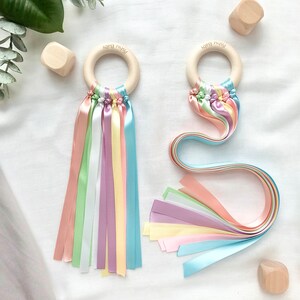 Puede incluir: Dos sonajeros de beb&eacute; de madera con cintas de colores pastel. Cada sonajero tiene un anillo de madera con el texto "Pishu Baby". Las cintas son en tonos pastel de rosa, verde, amarillo, morado y azul. Bloques de madera en el fondo.