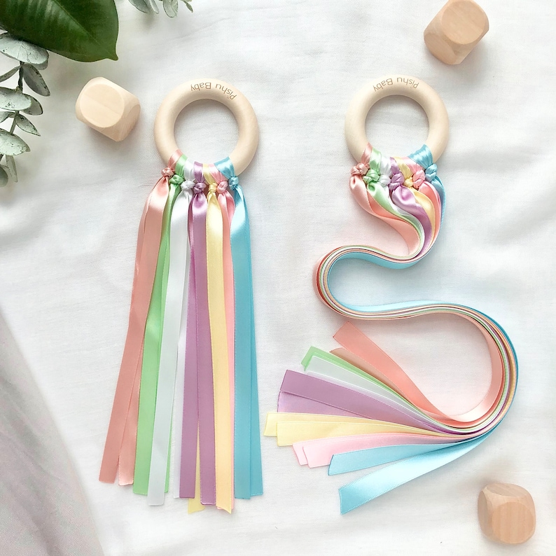 Puede incluir: Dos sonajeros de beb&eacute; de madera con cintas de colores pastel. Cada sonajero tiene un anillo de madera con el texto "Pishu Baby". Las cintas son en tonos pastel de rosa, verde, amarillo, morado y azul. Bloques de madera en el fondo.