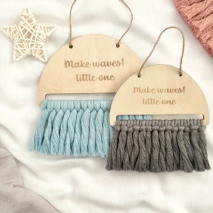 May include: Two wooden half-circle wall hangings with the text "Make waves! little one" and tassels. One hanging has light blue tassels and the other has gray tassels.