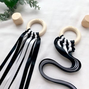 Puede incluir: Dos sonajeros de beb&eacute; de madera con cintas blancas y negras. Los sonajeros tienen un anillo de madera clara con el texto "Pishu Baby" impreso. Las cintas est&aacute;n unidas al anillo y cuelgan. Las cintas son blancas y negras.