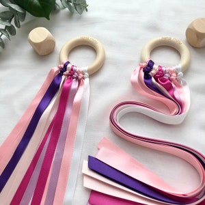 Puede incluir: Dos juguetes de madera para beb&eacute;s con un anillo de madera y cintas de colores en tonos rosa, morado y blanco. El texto "Pishu Baby" est&aacute; grabado en los anillos de madera. Tambi&eacute;n se ven cintas y bloques de madera adicionales.