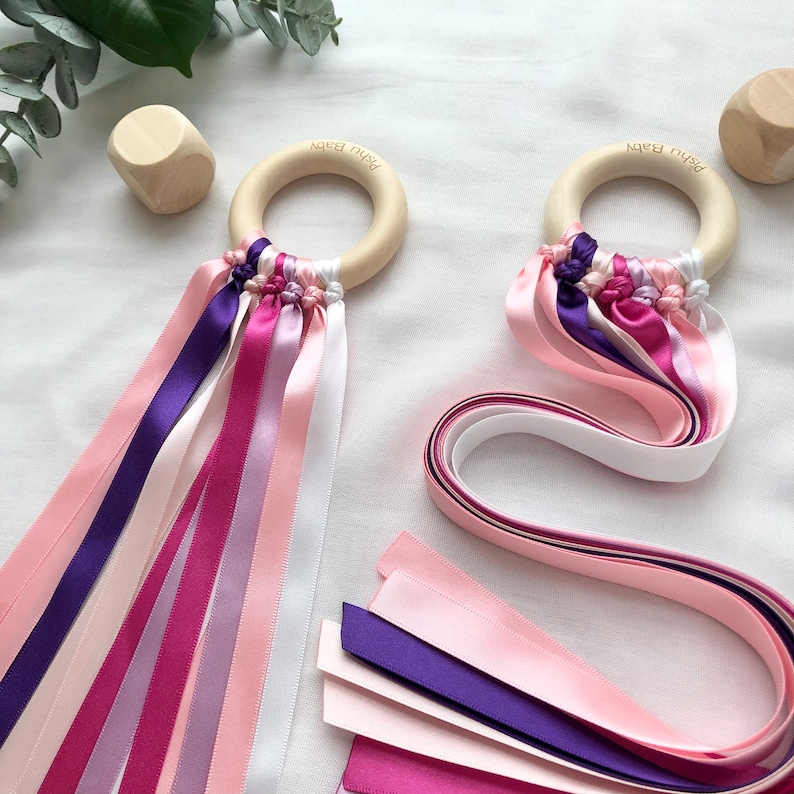 Puede incluir: Dos juguetes de madera para beb&eacute;s con un anillo de madera y cintas de colores en tonos rosa, morado y blanco. El texto "Pishu Baby" est&aacute; grabado en los anillos de madera. Tambi&eacute;n se ven cintas y bloques de madera adicionales.