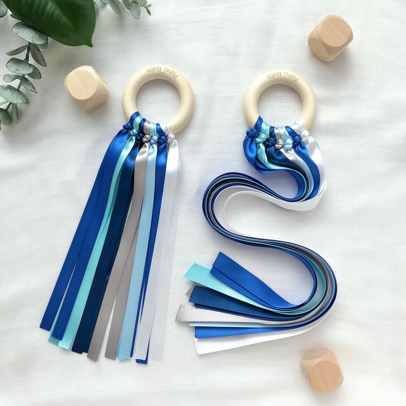 Puede incluir: Dos anillos de dentici&oacute;n de madera para beb&eacute;s con cintas de sat&eacute;n azules, plateadas y blancas. Los anillos tienen el texto "Pishu Baby" grabado. Varios cubos de madera peque&ntilde;os est&aacute;n esparcidos alrededor de los anillos.