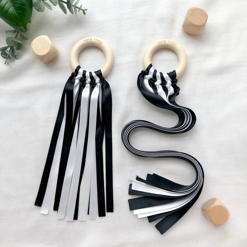 Puede incluir: Dos juguetes sensoriales para beb&eacute;s con aros de madera y cintas blancas y negras. Los aros tienen grabado el texto "Pishu Baby". Las cintas son una mezcla de blanco y negro. Tambi&eacute;n hay tres peque&ntilde;os bloques de madera.