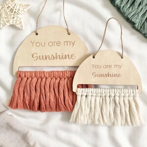 May include: Two wooden wall hangings with the text "You are my Sunshine" on each. The top hanging has a peach colored tassel fringe and the bottom hanging has a white tassel fringe.