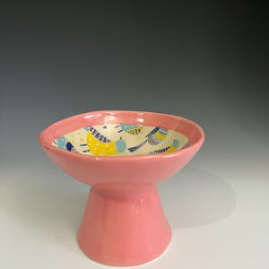 May include: A pink ceramic pedestal bowl with a wide rim and a flared base. The interior of the bowl features a colorful pattern of birds in yellow, blue, and white against a cream background. The bowl is a decorative item.