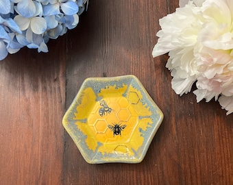 Puede incluir: Un pequeño plato hexagonal de cerámica con un esmalte amarillo y un borde azul. El plato presenta un patrón de panal con dos abejas negras.