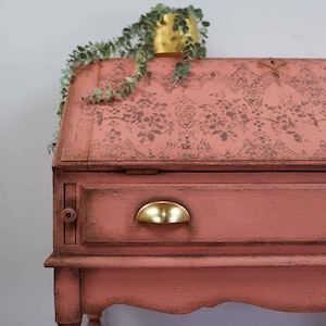 Op de afbeelding: Een koraalkleurig klepbureau met een donker bloemmotief op het schuine deksel, een enkele lade met een gepolijste goudkleurige halvemaanvormige handgreep en een kleine groene plant in een pot op de bovenkant. De tekst KACHA is zichtbaar.
