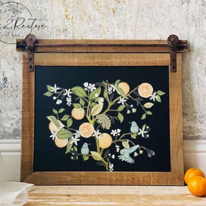 May include: A framed artwork featuring a black background with a botanical illustration of an orange tree with oranges, white blossoms, and small blue birds. The frame is made of light brown wood with rustic hardware.