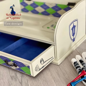 May include: Cream-colored wooden golf-themed storage box with a blue velvet-lined drawer. The box features a blue and green argyle pattern, golf club illustrations, and a blue trophy emblem. A golf ball, glove, and tees are next to the box.