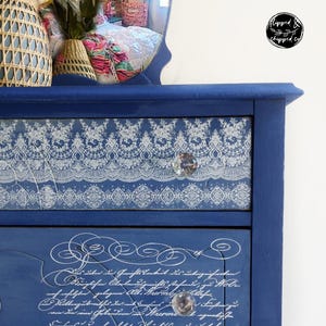 May include: A navy blue dresser with three drawers. The top drawer features a white lace design, and the middle drawer has script writing. Each drawer has a crystal knob. Floral accents adorn the bottom drawer.