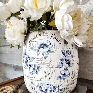 May include: A decorative vase with a distressed white finish, adorned with blue floral patterns and the text "MAISON DE PARIS." The vase is filled with large, white peony flowers. A wooden bead garland and a book are visible.