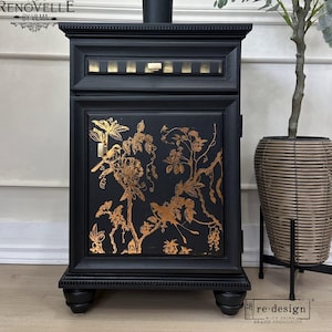 May include: Black cabinet with gold floral and bird design. The cabinet has a rectangular shape with a decorative top and bottom. The cabinet door features a detailed gold design of flowers, birds, and branches. The cabinet is on a light wood floor.