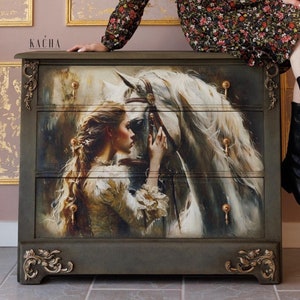 May include: A dark gray dresser with three drawers, featuring a painted image of a woman and a white horse. The dresser has ornate gold-colored accents and drawer pulls. The word "KACHA" is printed on the dresser.