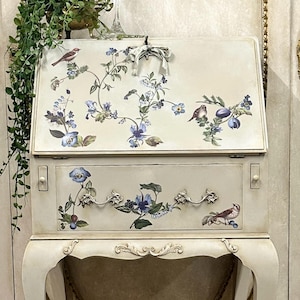 May include: An antique off-white secretary desk with floral and bird decals. The desk features a drop-down writing surface, a drawer, and curved legs. The desk has ornate hardware and a decorative chain. The text "re-design with prima" is visible.