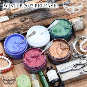 May include: Five small jars of metallic paint in various colors, including blue, white, red, gold, and green. The jars are arranged in a semi-circle on a wooden surface. The jars are labeled with the brand name "Finnabair".