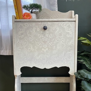 May include: A white painted wooden cabinet with a drop-down door and a shelf. The cabinet has a decorative scroll design and a black knob. The cabinet is sitting on a wooden floor with a plant in a wicker basket next to it.
