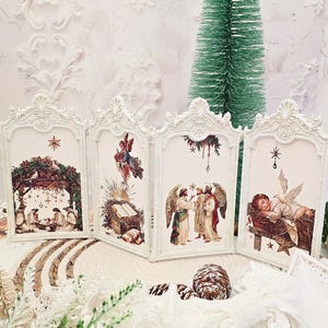 May include: A white, ornate, four-panel Christmas decoration featuring vintage-style illustrations of the Nativity scene. Each panel has a different image, including angels and the baby Jesus. A small green Christmas tree is in the background.