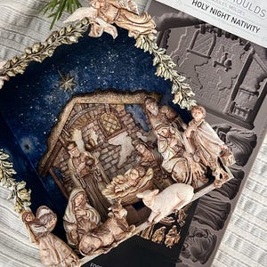 May include: A silicone mold for crafting, labeled Decor Moulds Holy Night Nativity, featuring detailed relief designs of a nativity scene with figures, an angel, and a stable. The flexible gray mold is displayed alongside a finished, painted nativity scene.