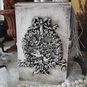 May include: A white wooden box with a silver-colored wreath design featuring two birds and a bow. The box has a distressed paint finish and a silver lid.