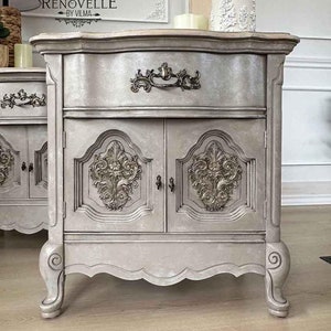 May include: A gray painted wooden cabinet with two doors and a drawer. The cabinet has ornate carvings on the doors and a silver-colored handle on the drawer. The cabinet is sitting on a wooden floor.