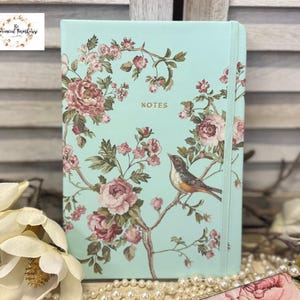 May include: A mint green notebook with a floral design featuring pink roses and a bird. The word "NOTES" is printed on the cover. A pearl necklace and white flowers are in the foreground.