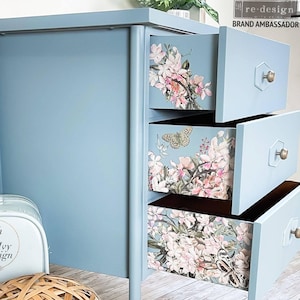 May include: A light blue painted desk with three drawers. The drawer fronts feature floral and butterfly designs. The desk has gold-colored hardware and is next to a small, white decorative box with the text "Blush & Ivy Design".