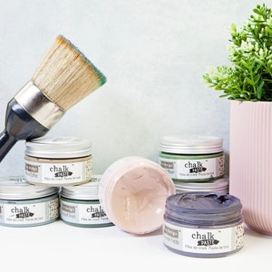 May include: Assortment of six small jars of chalk paste in various colors, including moss green, coffee, and gray. The jars are labeled "chalk paste" and have the text "Pâte de craie Pasta de tiza" printed on them. A paintbrush with green bristles is in the foreground.