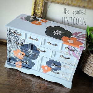 May include: A hand-painted jewelry box with a light blue base, adorned with floral designs in black, orange, and gray. The box features multiple drawers with ornate brass handles. The text "the painted UNICORN" is visible in the background.