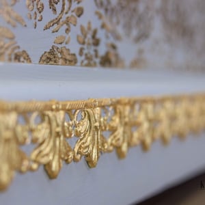 May include: Close-up of a white surface with a gold decorative trim. The trim is made of a delicate, intricate design with a floral motif.