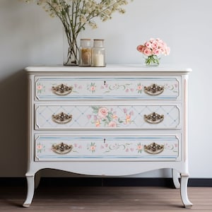 May include: A white dresser with three drawers features a hand-painted floral design with pink and blue accents. The dresser has ornate brass drawer pulls and sits on a wooden floor.