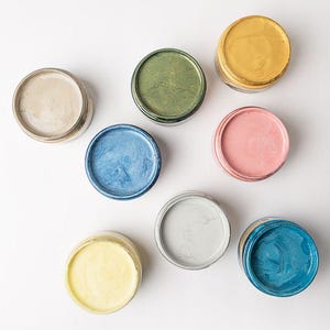 May include: Overhead shot of nine small, open jars of paint in various colors, including gold, blue, pink, and green. The jars are arranged on a white surface, showcasing the vibrant hues of the paint.