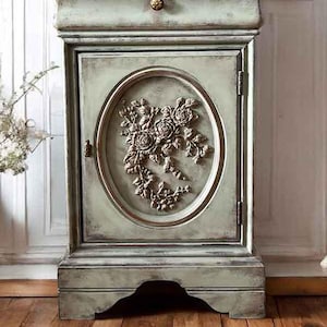 May include: A small, distressed green cabinet with a single drawer and a door featuring a floral design. The cabinet has a decorative base and is set against a white wood panel background.