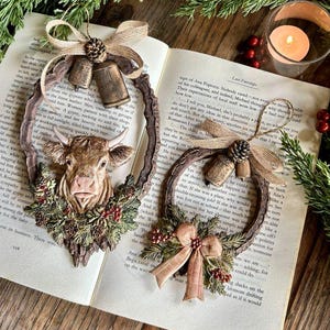 May include: Two oval-shaped hanging ornaments featuring a brown cow head, pinecones, red berries, and burlap bows. One ornament includes two small metal bells at the top, while the other features a single bell and a fabric bow at the bottom.