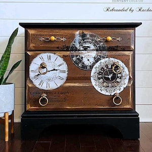May include: A wooden chest of drawers with a dark brown finish, featuring three drawers adorned with clock face decals. The top drawer has gold knobs, while the lower drawers have gold ring pulls. The chest sits on a black base.