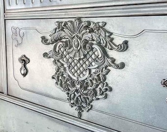 May include: A light blue antique buffet with ornate carvings and decorative hardware. The buffet has a wooden top and legs, and features a large decorative emblem on the front. The text "Dressers and Jujubes" and "re design" are visible.