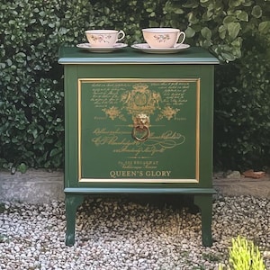 May include: Two green side tables with gold trim and floral designs, each topped with a teapot, teacups, and saucers. The tables are set against a backdrop of lush greenery and pink roses. The text "QUEEN'S GLORY" is visible.