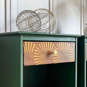 May include: Two green nightstands with gold-colored legs and drawer pulls. The drawer fronts feature a sunburst design in gold and brown. Decorative wire spheres sit atop one nightstand. The Grandson's Brush re-design logo is visible on the right nightstand.