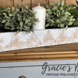 Op de afbeelding: Een lange, rechthoekige witte houten deegkom met een verweerde afwerking en een decoratief beige kantachtig patroon op de voorzijde. De kom bevat vier kleine groene potplanten en een enkele witte stompkaars.