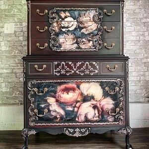 May include: An antique two-piece dresser in a dark finish, adorned with floral artwork on the drawers. It features ornate gold-coloured hardware and decorative trim. The top section has three drawers, and the bottom section has two drawers.