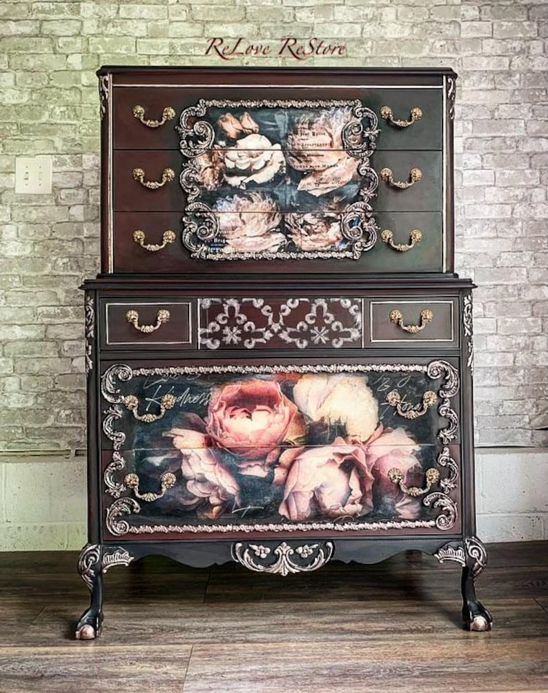 May include: An antique two-piece dresser in a dark finish, adorned with floral artwork on the drawers. It features ornate gold-coloured hardware and decorative trim. The top section has three drawers, and the bottom section has two drawers.