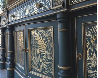 May include: A dark teal cabinet with gold accents and botanical designs. The cabinet features drawers, doors, and decorative columns. Gold botanical patterns adorn the doors and drawer fronts, creating an elegant and detailed look. The cabinet's trim is also gold.